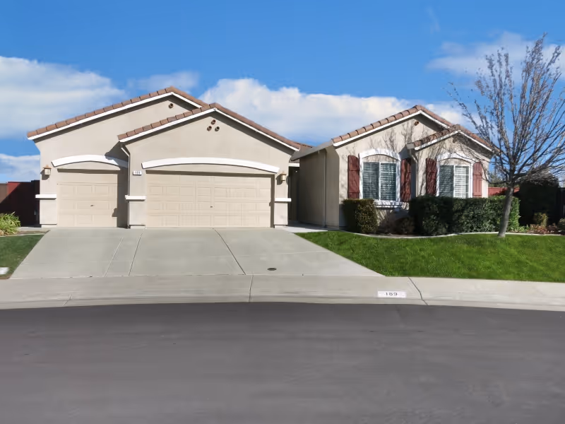 Exterior view of a single-story residential building with beige walls, three garage doors, a driveway, green lawn, and a tree on the right side under a blue sky with some clouds.