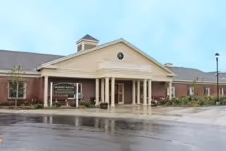 Exterior view of a single-story brick building with a covered entrance supported by columns. The building has a triangular pediment with a small circular window and a cupola on the roof. There is a sign near the entrance that reads 'Memory Care at Market Square'. The sky is overcast and the ground appears wet from rain.