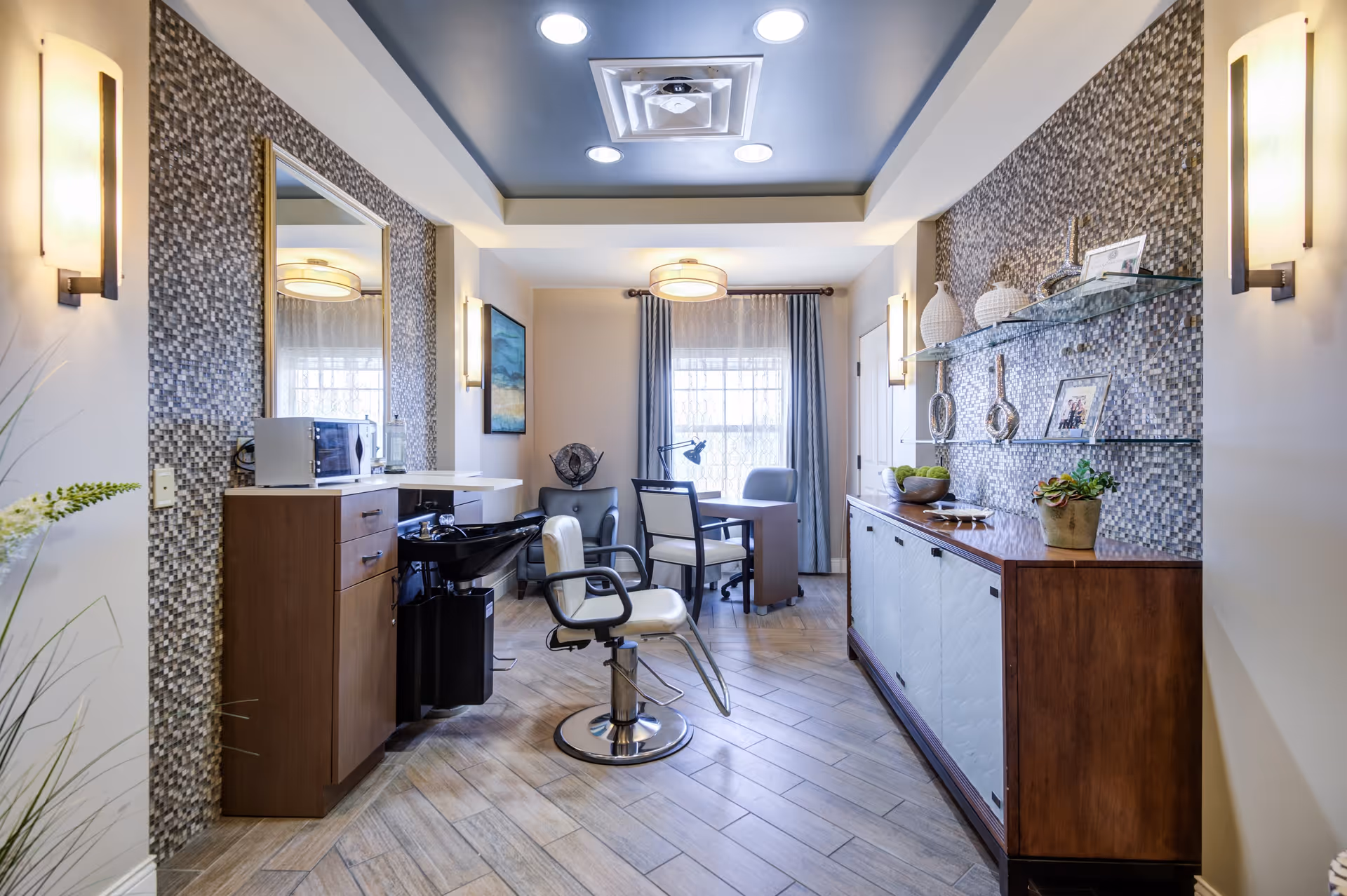 A modern salon area inside a senior living facility with a styling chair in front of a sink and mirror, wooden cabinets, decorative wall tiles, and a small table with chairs near a window with curtains.