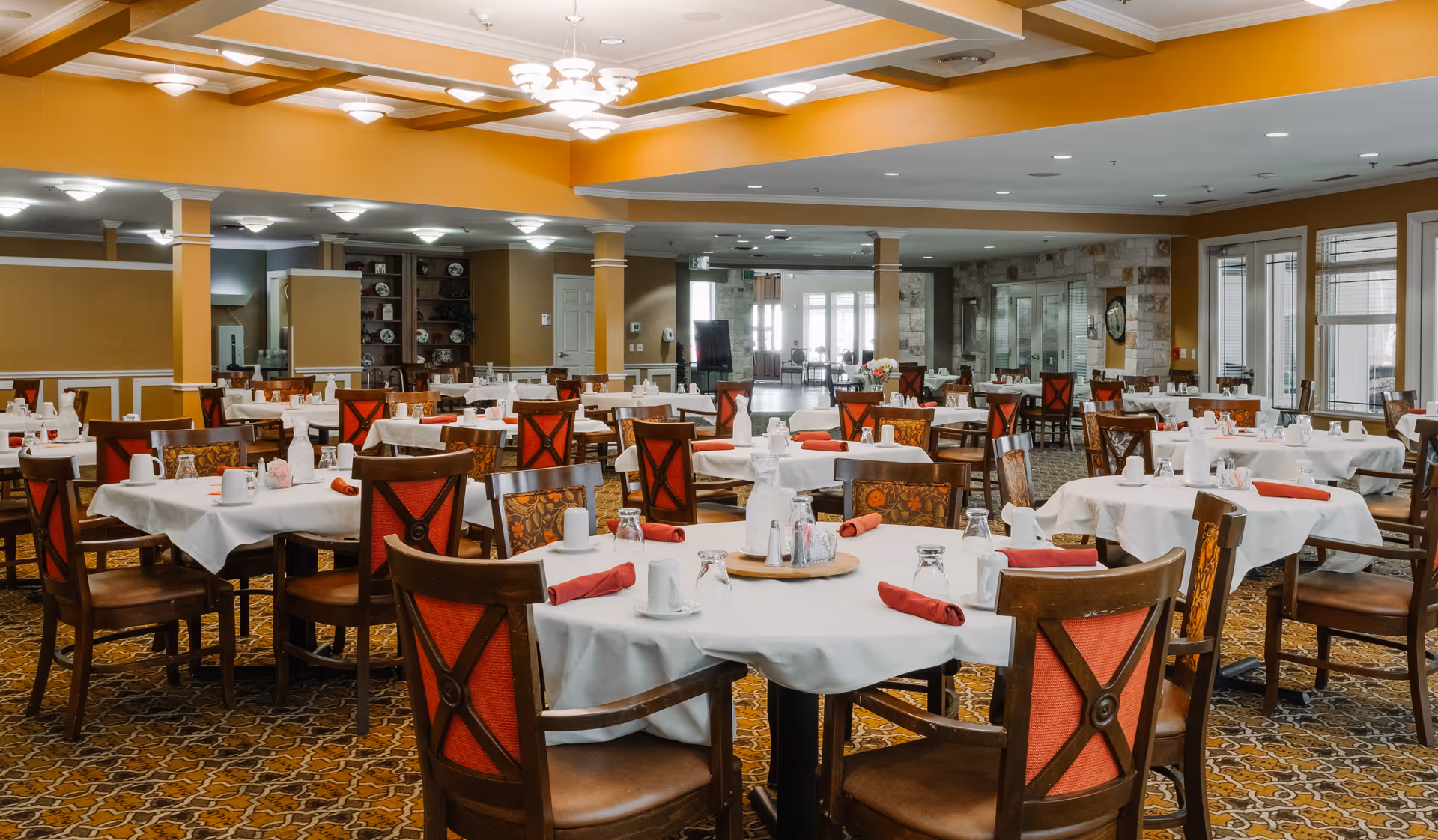 Spacious dining room with multiple round tables covered with white tablecloths, each set with glasses, cups, napkins, and condiments. The chairs have wooden frames with red and floral upholstery. The room features warm yellow walls, patterned carpet, and ceiling lights.