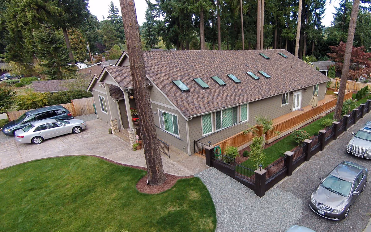 Aerial view of a single-story residential building with a brown shingle roof and multiple skylights. The building is surrounded by a well-maintained lawn, trees, and a fenced garden area. Several cars are parked in the driveway and along the adjacent gravel road. The setting is peaceful with tall pine trees in the background.