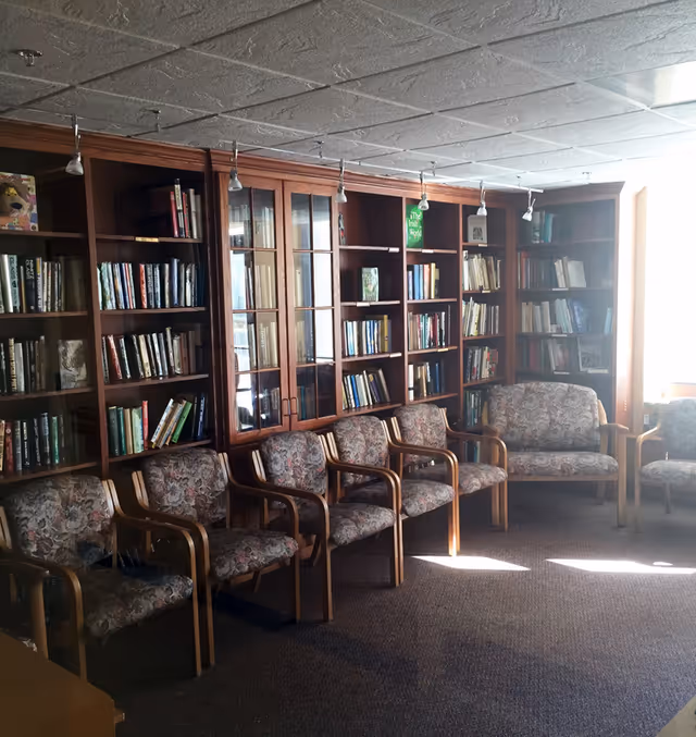 Wood-paneled library room with bookshelves and a row of upholstered chairs arranged along the wall.