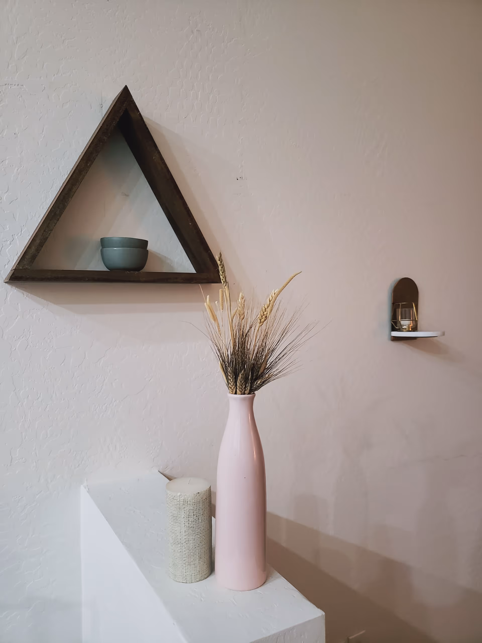 Interior wall with a triangular wooden shelf holding a small bowl, a tall pink vase of dried grasses on a pedestal, and a small wall shelf with a candle.
