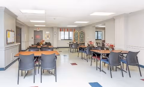 A bright dining room with several tables and chairs arranged neatly. The room has white walls with wainscoting, a bulletin board on the left wall, and a window with a striped valance at the far end. There are floral centerpieces on some tables and a decorative folding screen near the window.