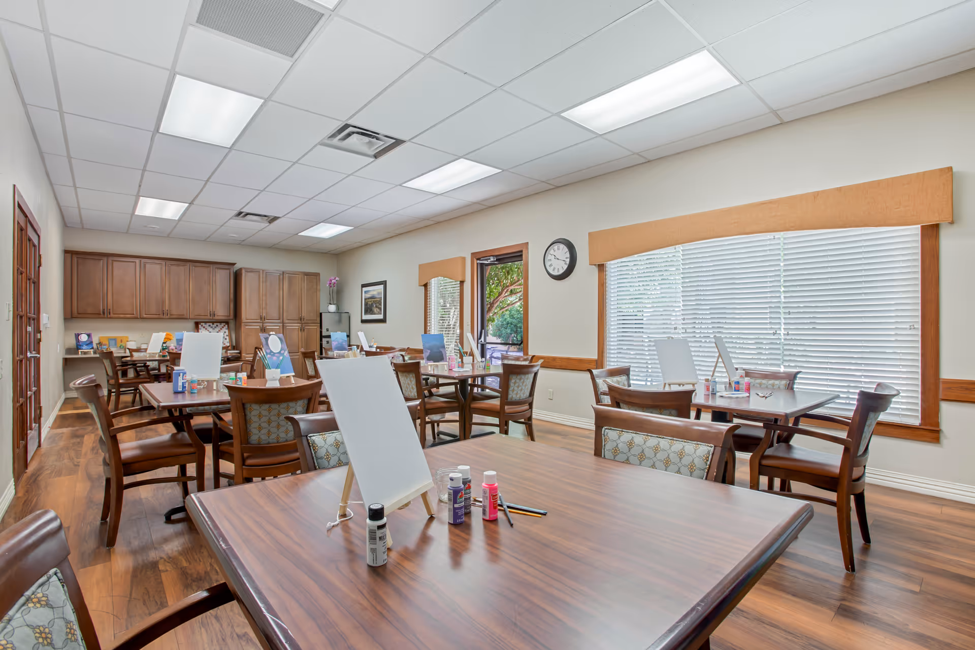 A bright activity room with several wooden tables and chairs arranged neatly. Each table has a small easel with a blank canvas and various paint bottles and brushes, suggesting a painting or art activity setup. The room has large windows with blinds, wooden flooring, and cabinets along one wall. A clock is mounted on the wall near the windows.