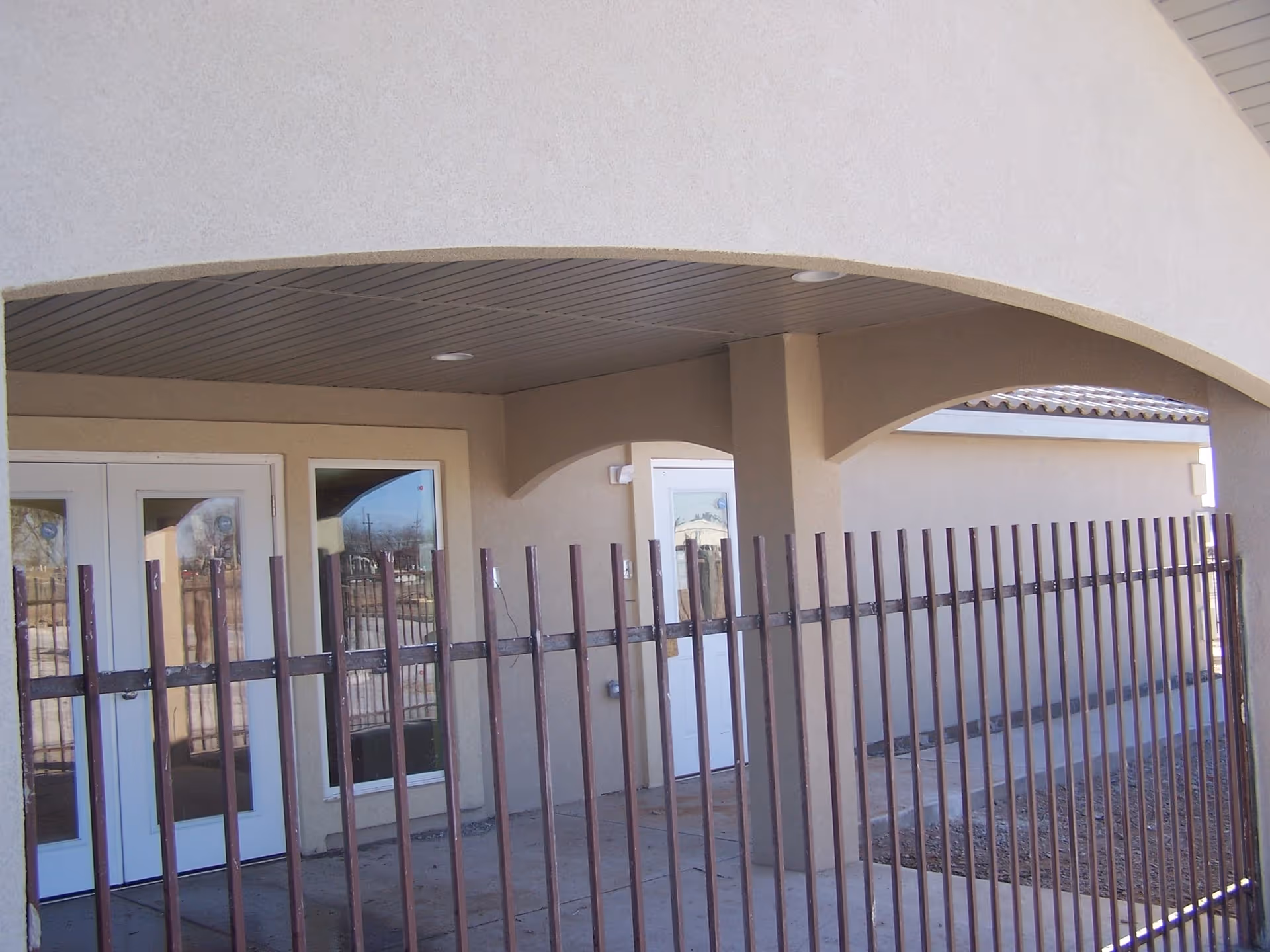 Exterior view of a senior living facility showing a beige stucco building with arched openings, white doors with glass panels, and a metal fence in the foreground.
