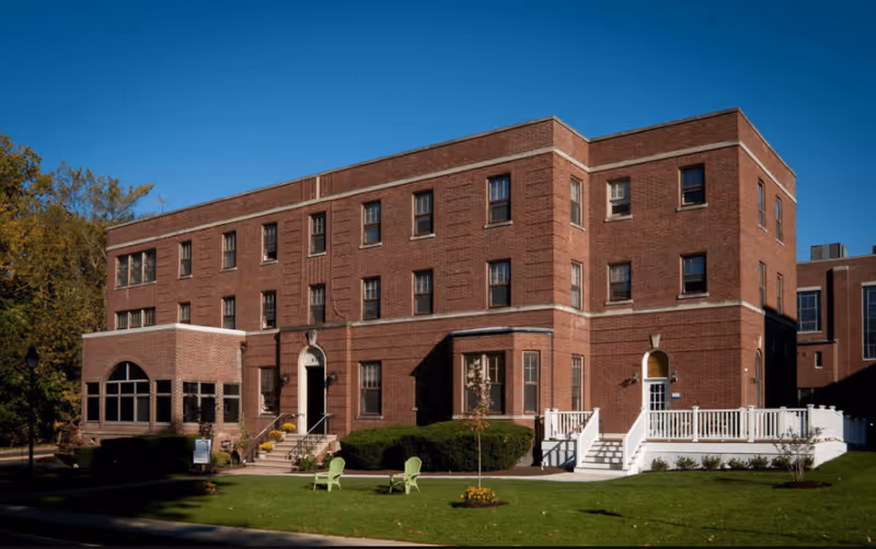 A large three-story brick building with multiple windows, a small porch with stairs, and a white railing on the right side. The building is surrounded by green grass, a few small trees, and two green chairs on the lawn. The sky is clear and blue.
