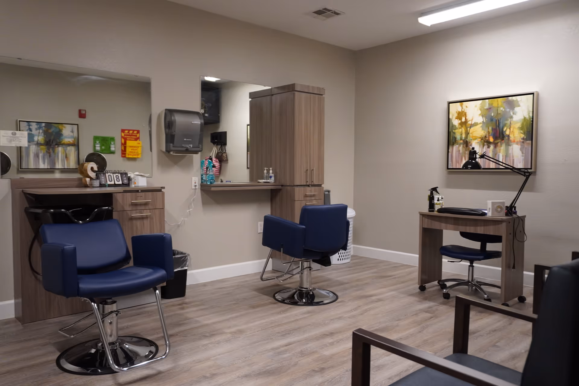 Interior of a salon area in a senior living facility with two blue salon chairs, a wooden cabinet, a mirror, a desk with a lamp, and a colorful abstract painting on the wall.