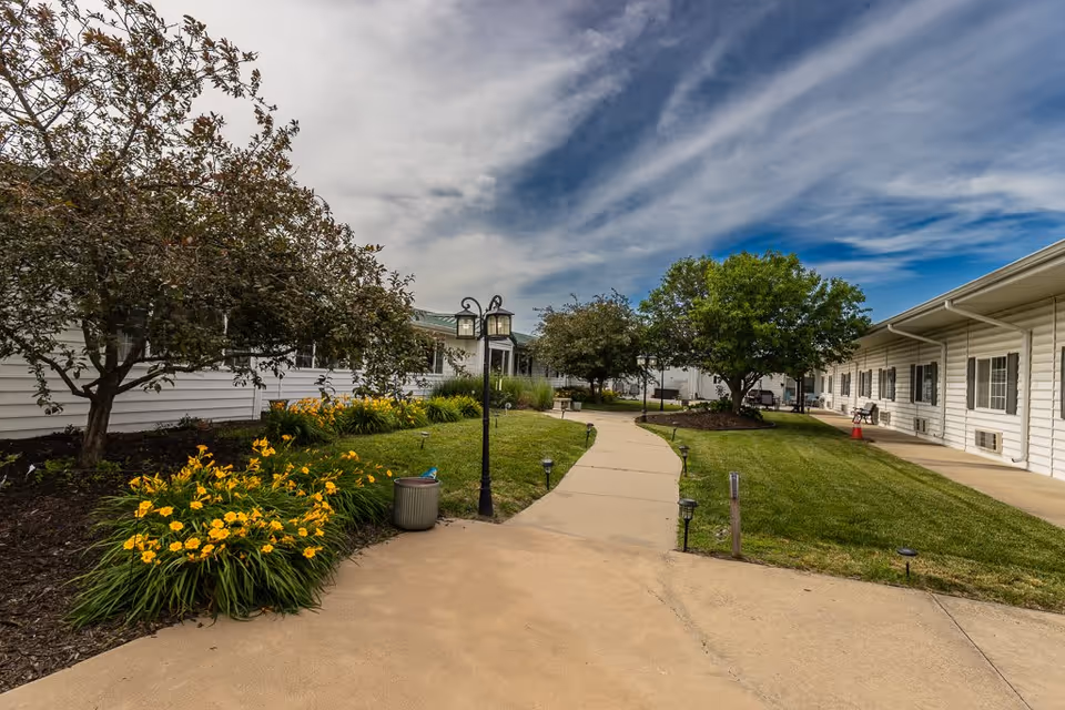 Outdoor walkway at Bailey Pointe at Plattsmouth with green grass, yellow flowers, trees, and white buildings on either side under a partly cloudy sky.