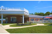 Single-story brick senior care facility with a covered entrance, paved walkways, and a grassy landscaped lawn under a clear blue sky.