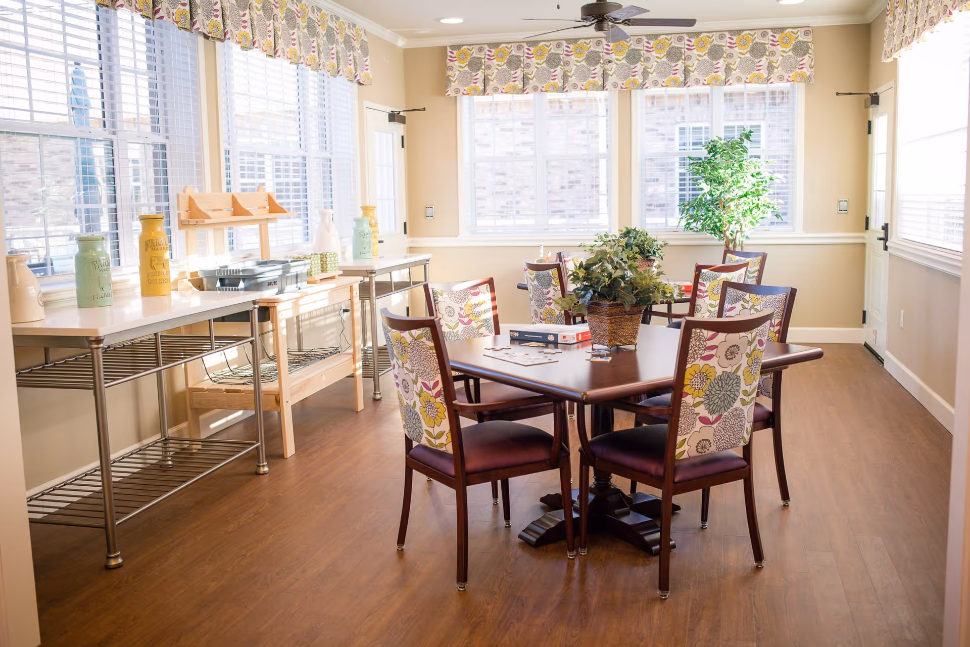 A bright and cozy dining area with a wooden table surrounded by six chairs featuring floral patterned upholstery. The room has large windows with floral valances allowing natural light to fill the space. There are decorative plants on the table and in the corner near the windows. Along one wall, there are metal and wooden shelves with decorative jars and containers. The floor is wooden, and a ceiling fan is mounted above.