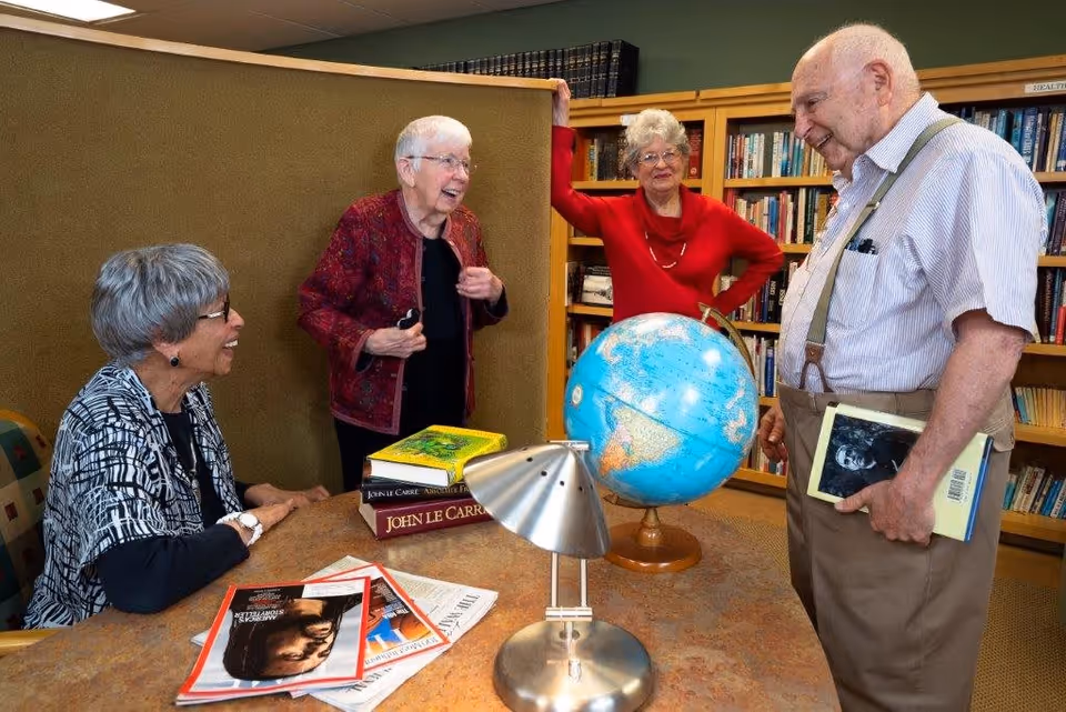 Four elderly people gathered around a table in a library or reading room. Three women and one man are engaged in conversation. On the table are several books, magazines, and a globe. Bookshelves filled with books are visible in the background.