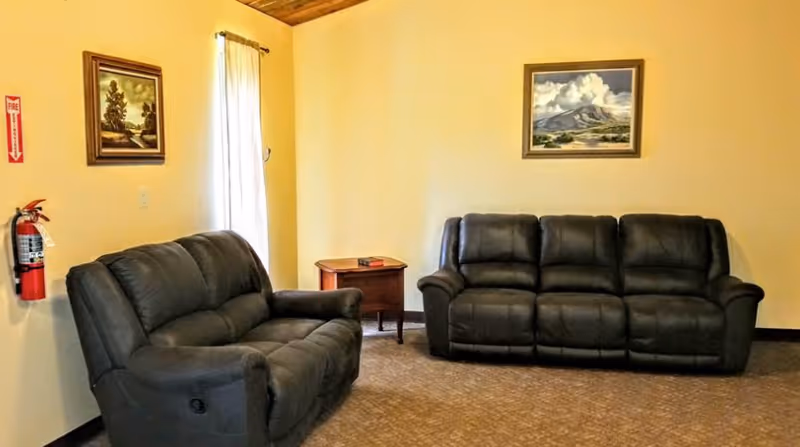 A cozy living room with two dark gray reclining sofas, a small wooden side table with a book on it, beige walls, a window with a curtain, and two framed landscape paintings hanging on the walls. A fire extinguisher is mounted on the left wall.