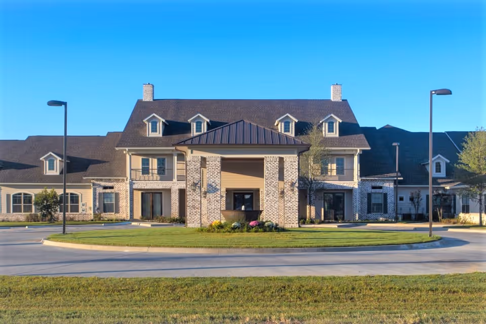 Front exterior view of The Claiborne at Thibodaux, a large two-story building with a peaked roof, multiple dormer windows, and a covered entrance supported by brick columns. The building is surrounded by a circular driveway, street lamps, and well-maintained grass and landscaping under a clear blue sky.