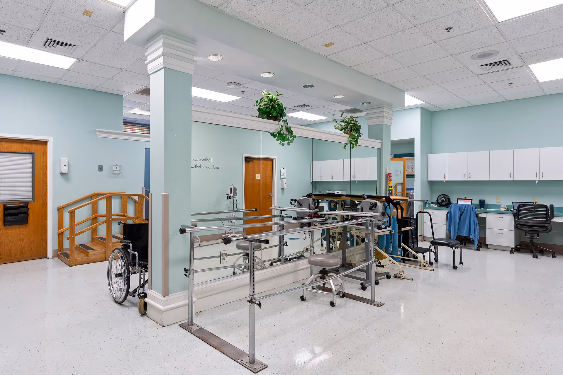 A rehabilitation or physical therapy room with parallel bars for walking exercises, a wheelchair, a small set of stairs with a handrail, chairs, desks with office supplies, and cabinets. The walls are painted light blue, and there are plants hanging from the ceiling. The room is well-lit with fluorescent lights.