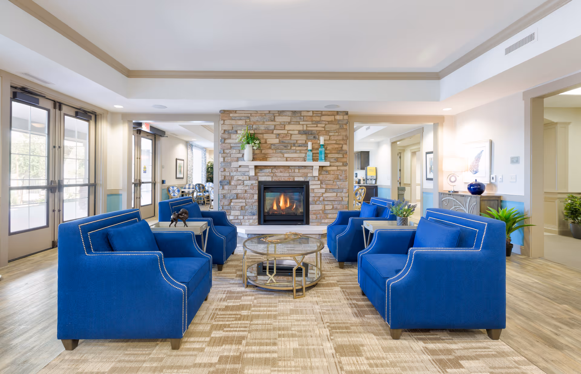 A bright and spacious senior living facility lounge area with four blue upholstered armchairs arranged around a round glass coffee table. A stone fireplace with decorative vases and plants is centered on the far wall. Large windows and glass doors let in natural light, and the room features light-colored walls and wood flooring.