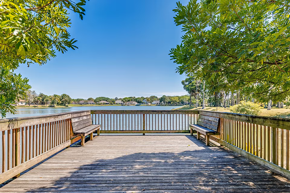 Wooden deck with benches on both sides overlooking a calm lake, surrounded by trees and houses in the distance under a clear blue sky.