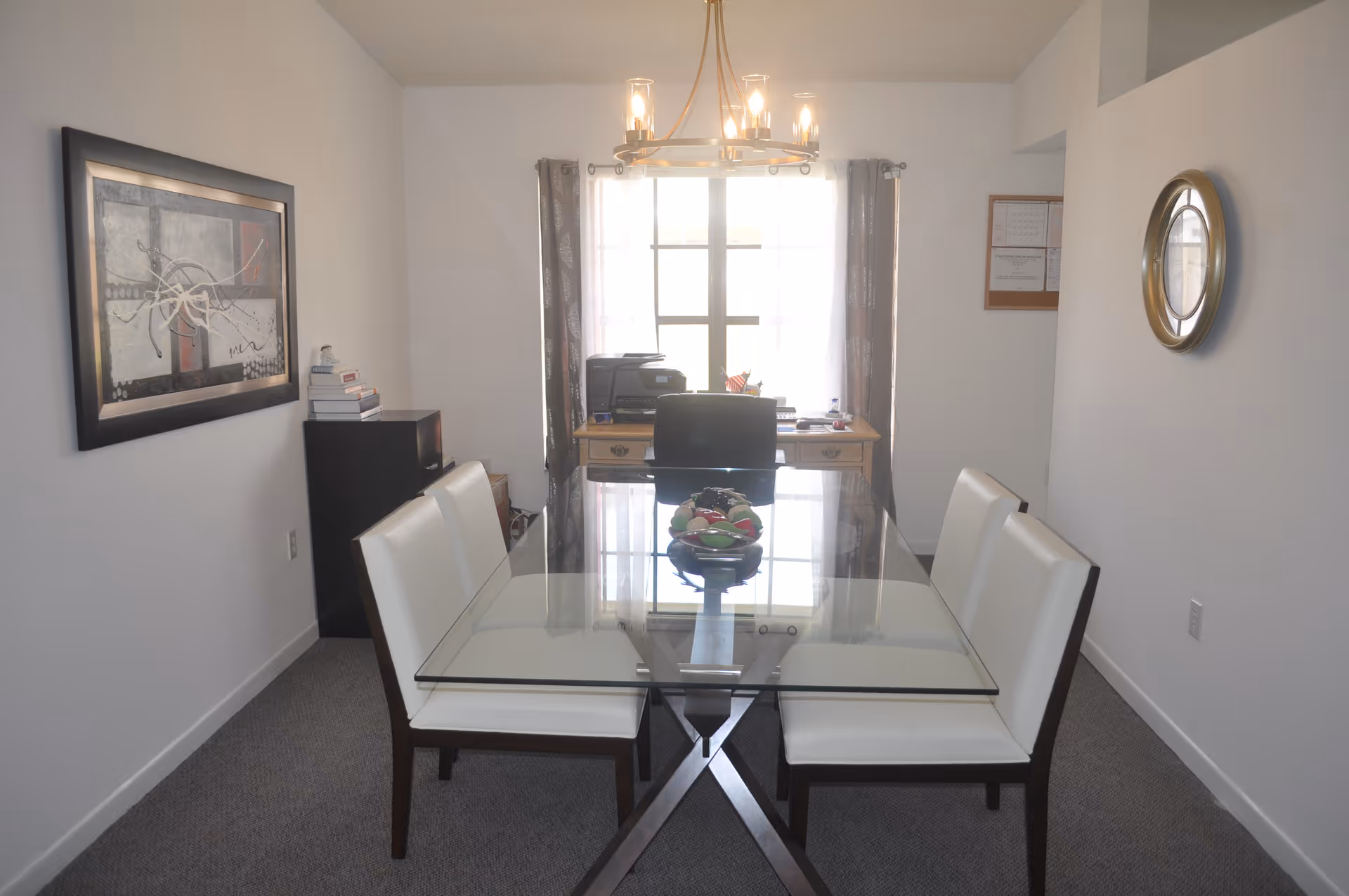 A dining room with a glass-top table and four white cushioned chairs. At the far end, there is a wooden desk with a chair, a printer, and some office supplies in front of a window with curtains. The walls are white, with a framed abstract painting on the left wall and a round clock on the right wall. A modern chandelier hangs above the table.