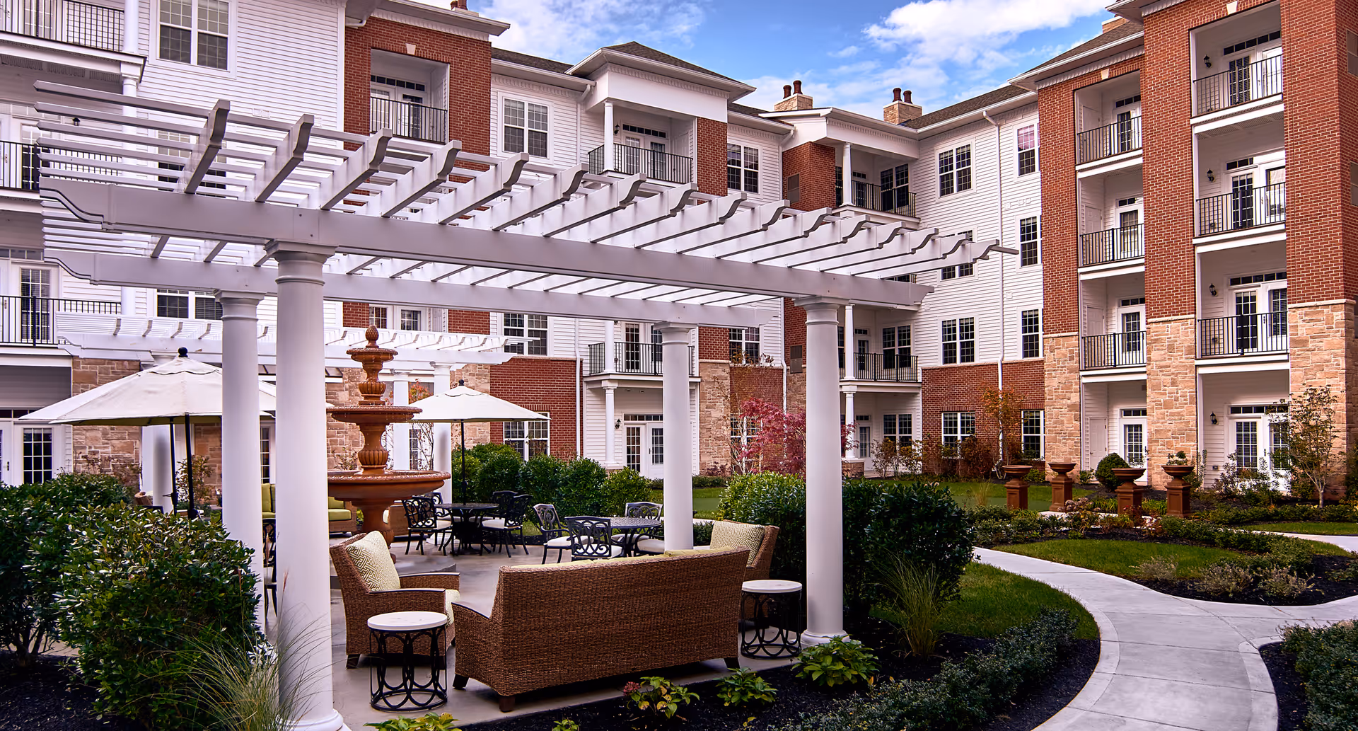 Outdoor seating area at Waltonwood at Ashburn featuring wicker furniture with cushions under a white pergola, surrounded by greenery and a multi-tiered fountain. The background shows a multi-story residential building with balconies and large windows.