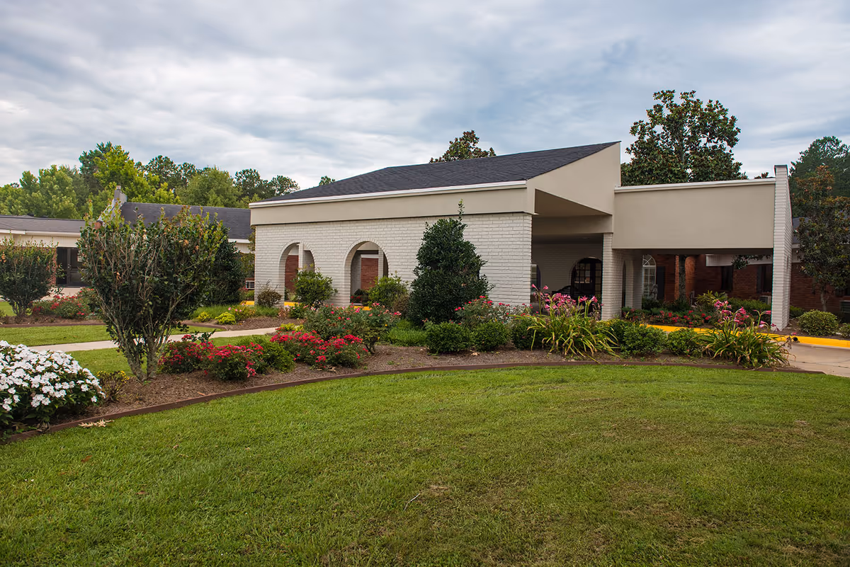 Front entrance of a single-story care facility with a covered porte-cochere, manicured lawn and flower beds.