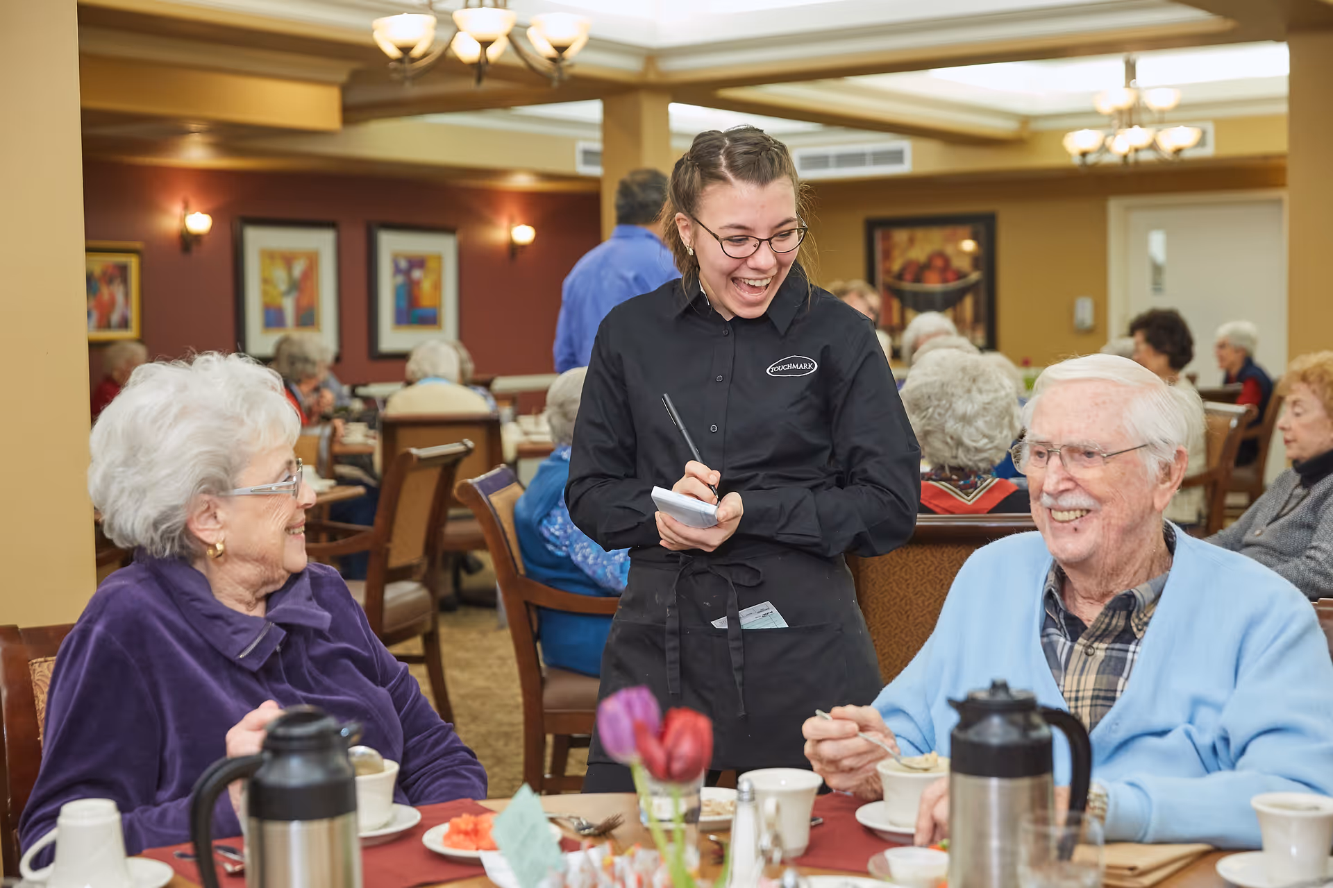 A smiling waitress wearing a black uniform takes an order from two elderly people seated at a dining table in a busy dining room. The elderly woman on the left wears a purple jacket and glasses, and the elderly man on the right wears a light blue cardigan and glasses. The table has coffee pots, cups, and a small vase with flowers. Other elderly people are seated and dining in the background.
