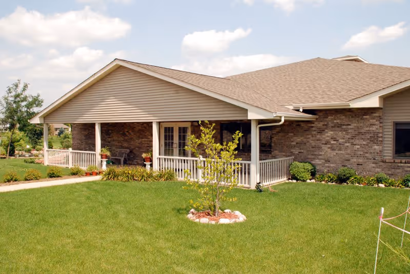 Single-story brick building with a covered porch, white railing, and a small tree planted in a circular stone-bordered bed on a well-maintained green lawn under a partly cloudy sky.
