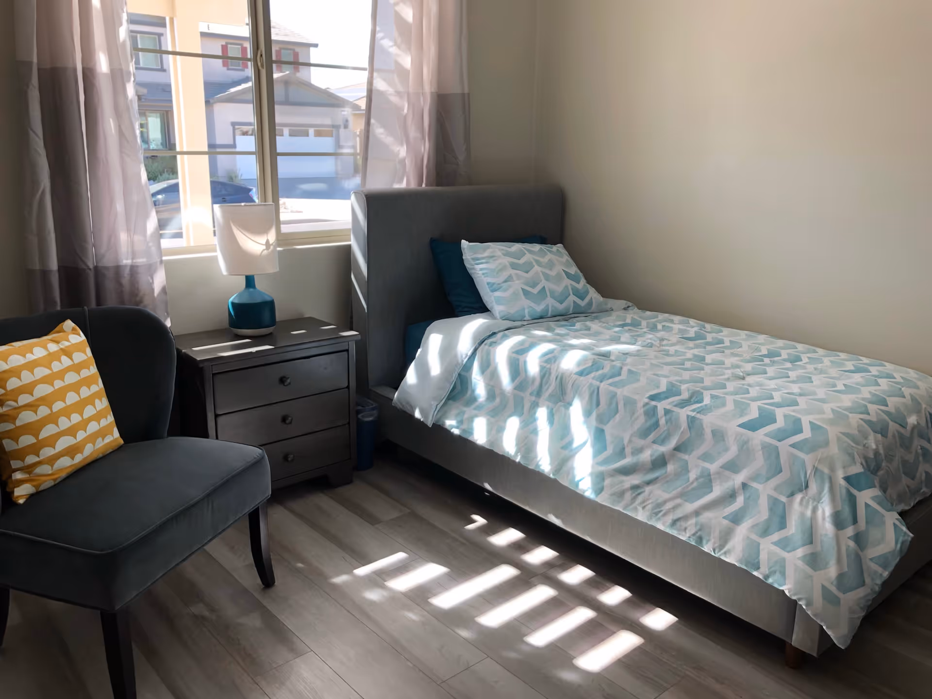 Sunlit single bedroom with a patterned bed, nightstand and lamp, and an upholstered chair by a window.
