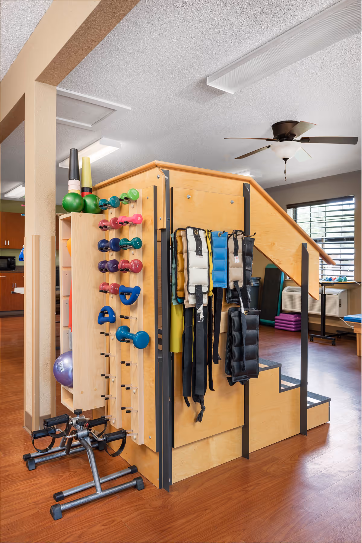 Exercise/physical therapy area with a wooden stair structure holding dumbbells, resistance weights, ankle weights, and a pedal exerciser.