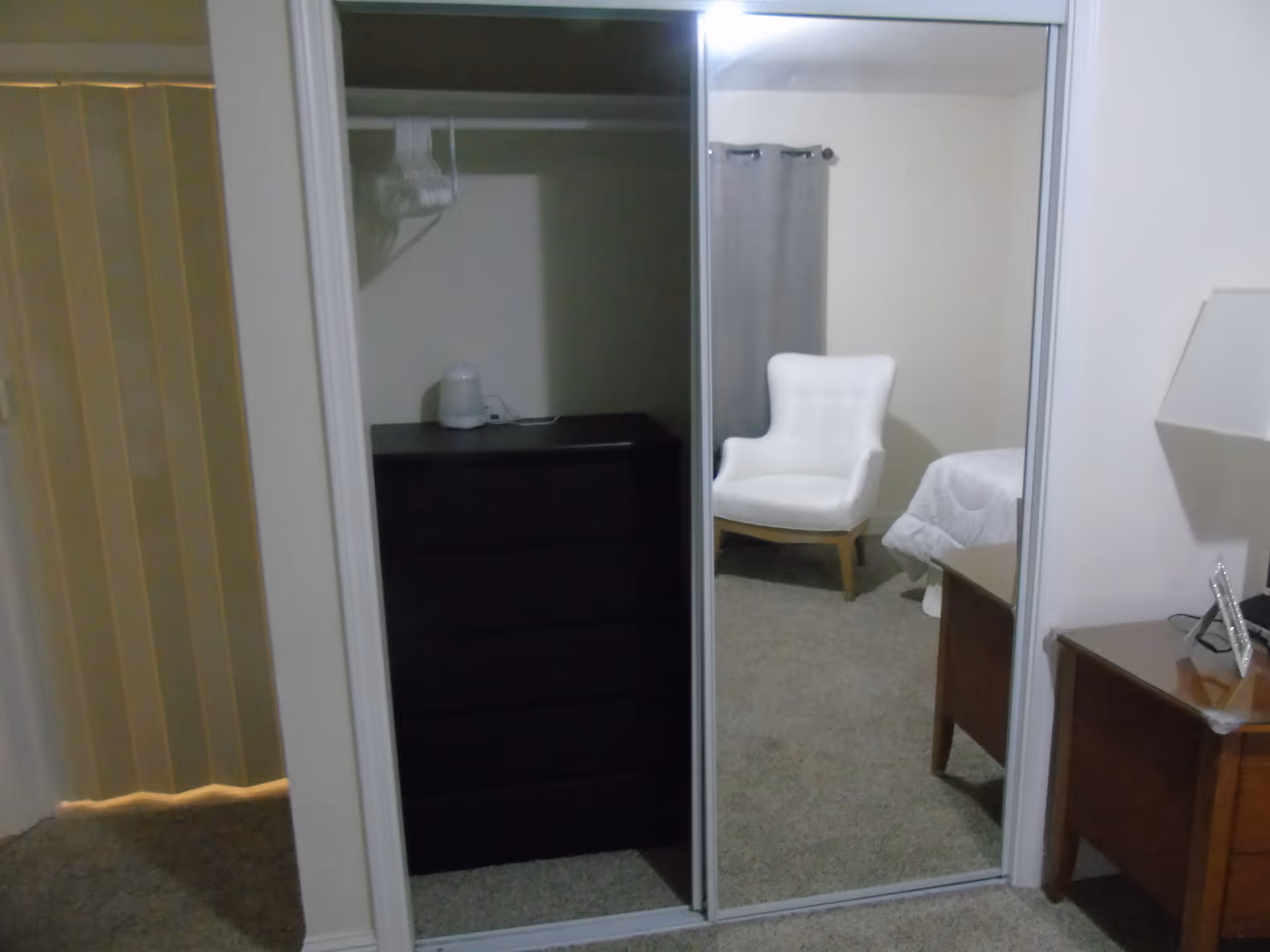 Mirrored closet doors reflecting a bedroom with a white armchair, dresser, nightstand with a lamp and part of a bed.