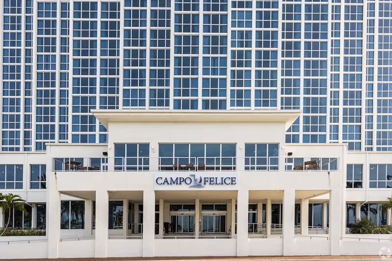 Front exterior view of a tall building with many windows and a white entrance structure displaying the sign 'Campo Felice'. There are outdoor seating areas on the balcony above the entrance.