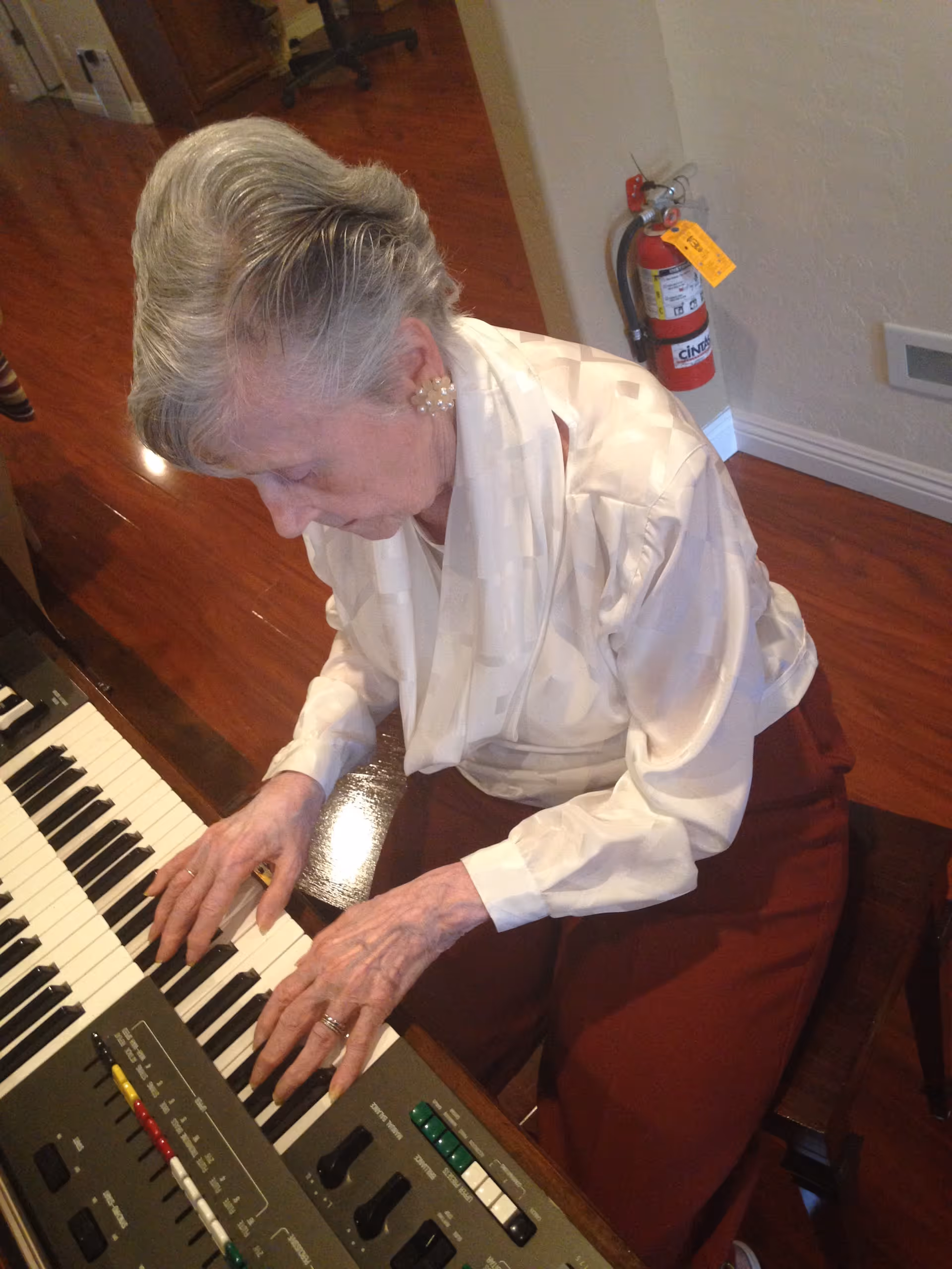 An elderly woman with gray hair styled neatly is playing an electronic keyboard piano indoors. She is wearing a white blouse and maroon pants, seated on a wooden chair on a polished wooden floor. A fire extinguisher is mounted on the wall behind her.