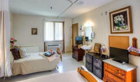 A furnished patient room with a bed, dresser, desk, and TV in a senior facility.
