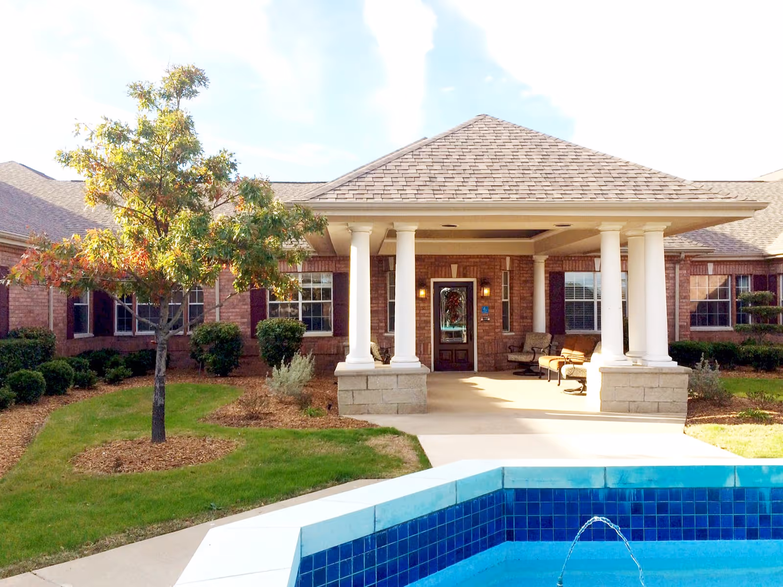 Exterior view of a brick building with a covered entrance supported by white columns. There is a small fountain with blue tiles in the foreground, a tree with green and orange leaves, and some shrubs and grass around the building.