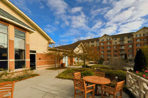 Courtyard patio with wooden tables and chairs in front of a multistory senior living building under a partly cloudy sky.