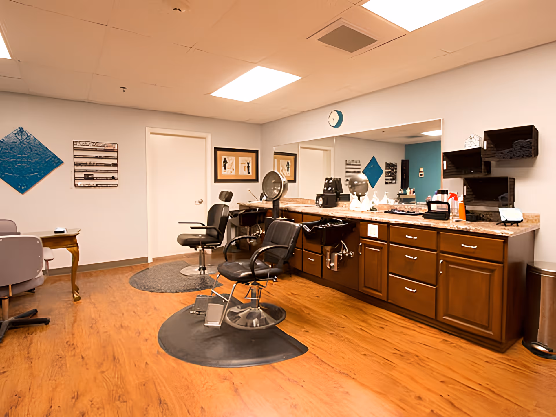 Interior view of a salon area in a senior living facility with two black salon chairs, a large mirror above a countertop with cabinets, and various hair styling tools and products. The room has wooden flooring, light-colored walls with some artwork, and a small table with chairs in the corner.