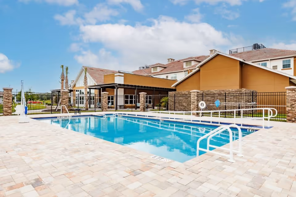 Outdoor swimming pool with clear blue water surrounded by a tiled deck. The pool area is enclosed by a black metal fence and has white handrails and a pool lift for accessibility. In the background, there are buildings with beige and brown exteriors under a partly cloudy blue sky.