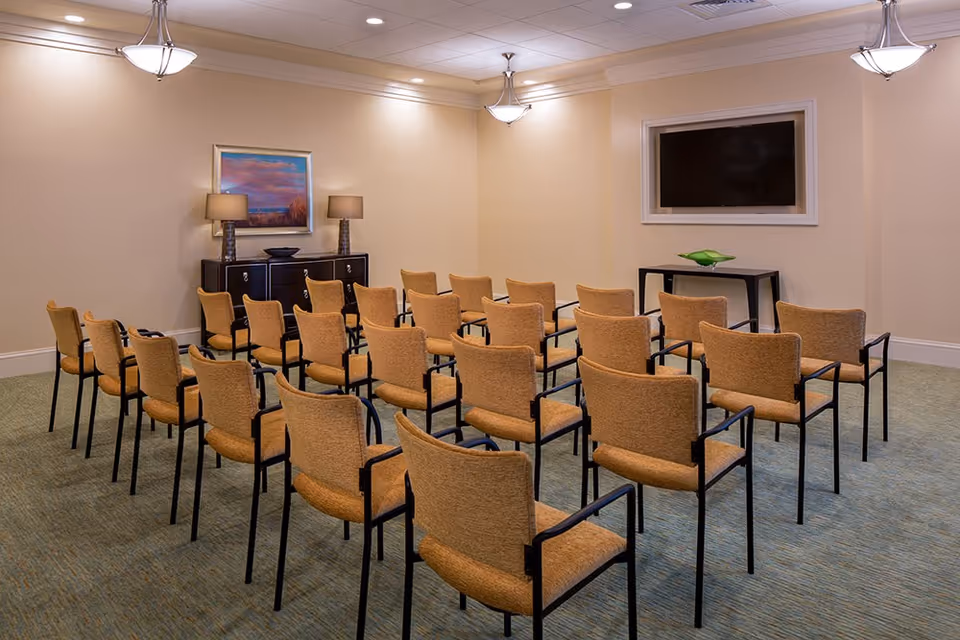 A small conference or meeting room with rows of beige upholstered chairs arranged facing a wall-mounted flat screen TV. The room has beige walls, carpeted floor, two table lamps on a dark wooden cabinet, a framed landscape painting, and ceiling light fixtures.
