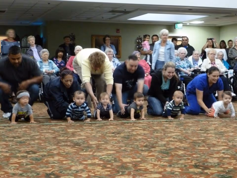 A group of babies crawling on a patterned carpet indoors, each accompanied by an adult. In the background, elderly people are seated and watching the activity in a room with green walls and ceiling lights.