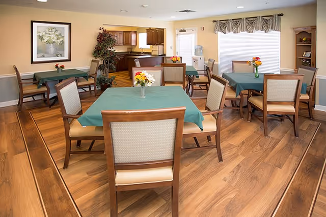 Dining room with several square tables covered in green tablecloths, wooden chairs, hardwood floors, floral centerpieces, and a kitchenette in the background.