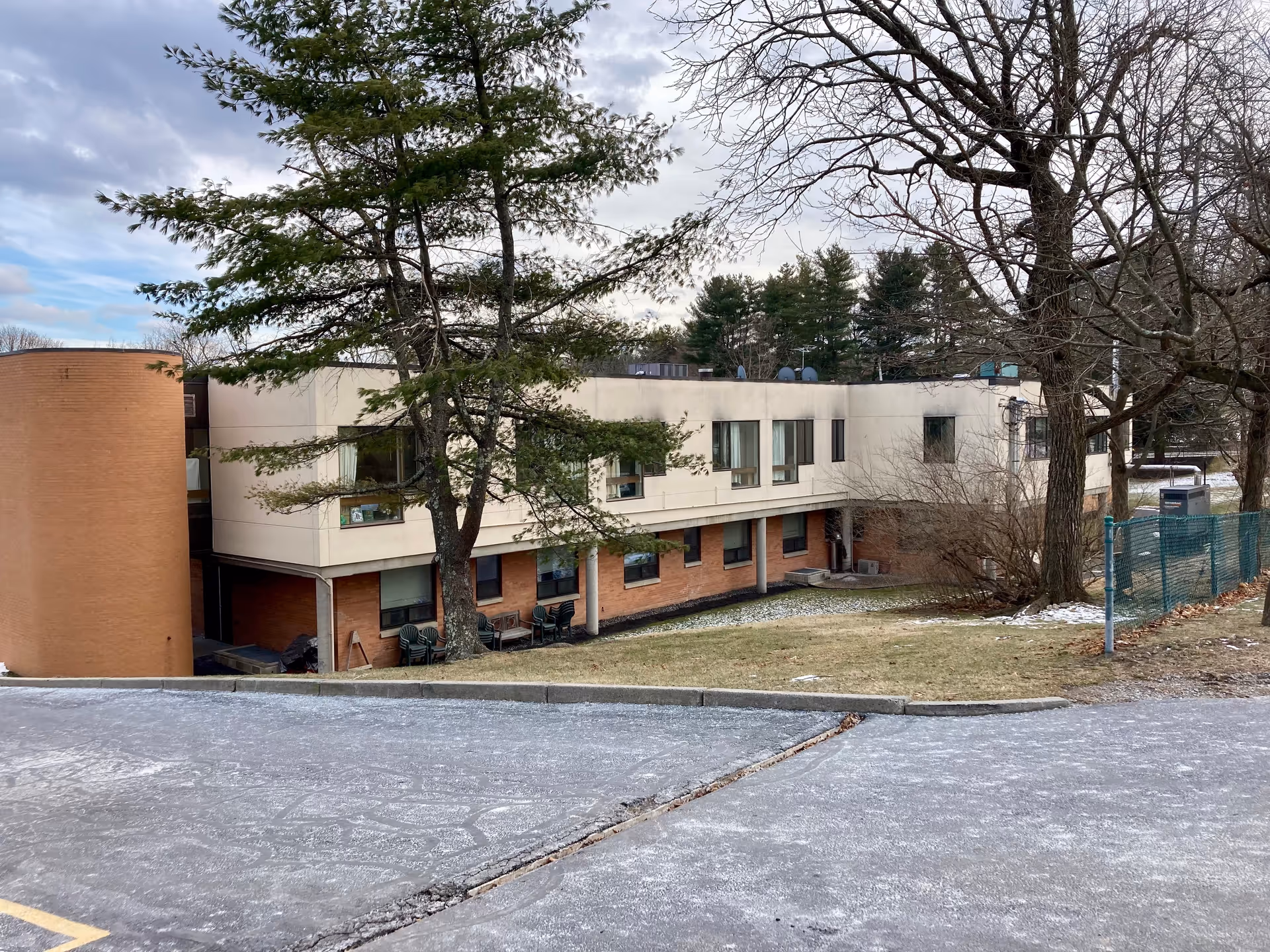 Exterior view of a two-story nursing and rehabilitation center building with a mix of brick and light-colored walls, surrounded by trees and a partially snow-covered lawn.