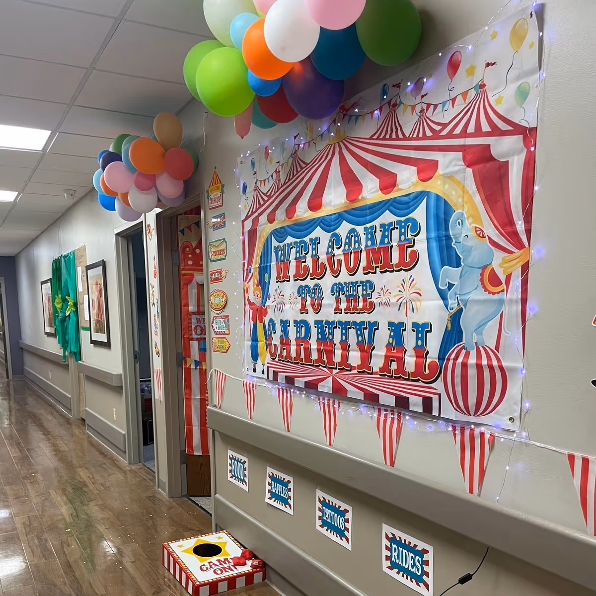 A decorated hallway in a skilled nursing and rehabilitation center with a carnival theme. The wall features a large colorful banner that reads 'Welcome to the Carnival' with circus tent and elephant illustrations. Above the banner, there are clusters of colorful balloons attached to the ceiling. The hallway has wooden floors, framed pictures on the walls, and carnival-themed signs and decorations, including a small game box on the floor labeled 'Game On!'.