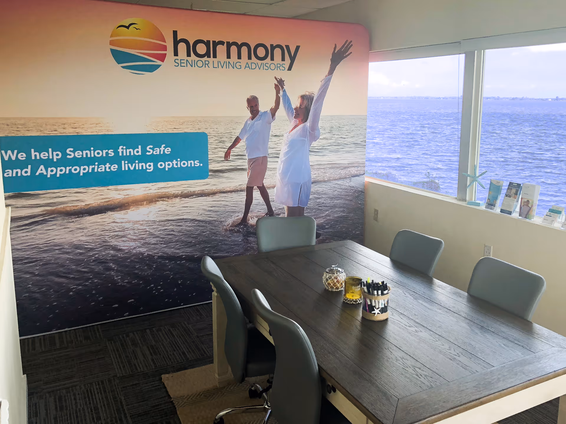 Small meeting room with a wooden table and chairs in front of a large 'Harmony Senior Living Advisors' beach mural and a window overlooking the sea.
