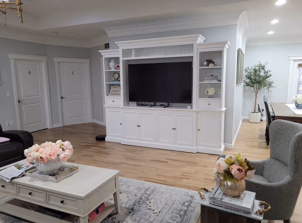 A cozy living room area with light wood flooring, a white entertainment center holding a large flat-screen TV, and two white doors labeled 4 and 5. The room features a white coffee table with a vase of pink flowers, a gray armchair with a side table holding another vase of flowers, and a dining area with a wooden table and chairs in the background. There is also a potted plant near the dining area.