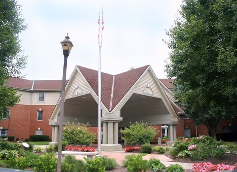 Covered porte-cochere entrance of a brick-and-siding building with an American flagpole and landscaped flower beds.