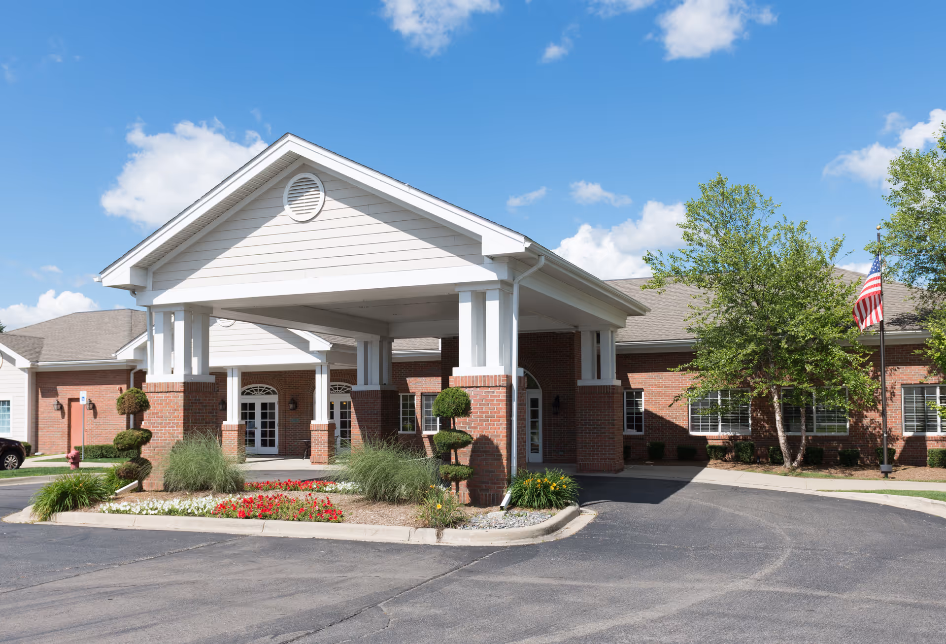 Front exterior view of a single-story brick building with a covered entrance supported by white columns. There are landscaped flower beds and trimmed bushes near the entrance, a paved driveway, and an American flag on a flagpole to the right. The sky is clear with a few clouds.