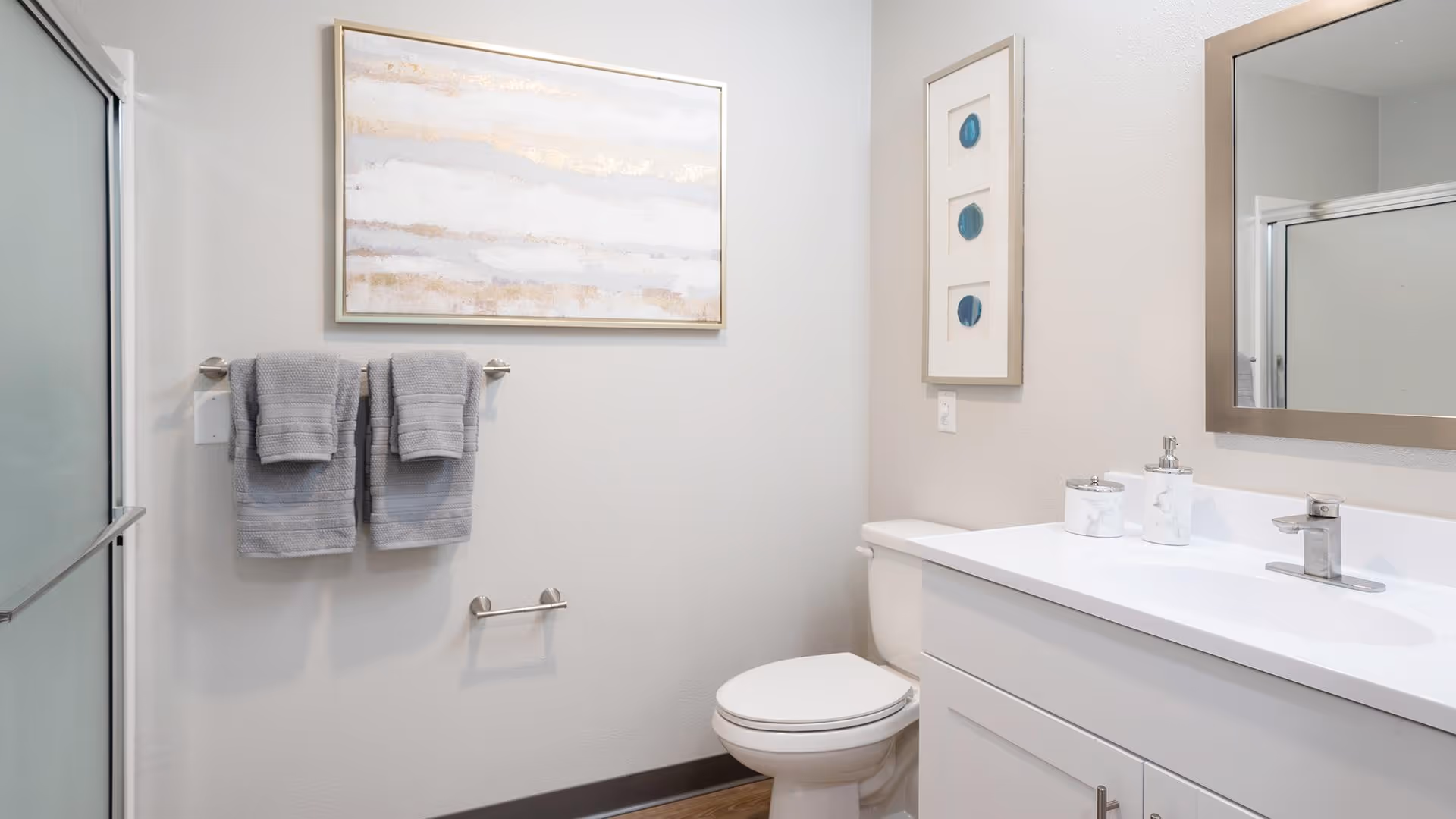 A clean and modern bathroom featuring a white toilet, a white vanity with a sink and silver faucet, a large mirror above the sink, two gray towels hanging on a towel rack, a glass shower door, and framed artwork on the walls.