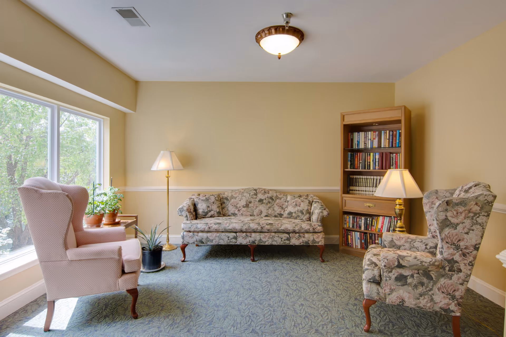 A cozy living room with floral patterned sofa and armchair, a pink upholstered wingback chair, a wooden bookshelf filled with books, two floor lamps, and several potted plants near a large window letting in natural light.