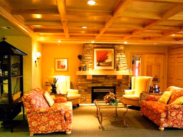 Cozy living room with floral patterned sofas, two beige armchairs, a glass coffee table with a flower arrangement, and a stone fireplace with a framed artwork above it. The room has warm lighting and wooden ceiling beams.
