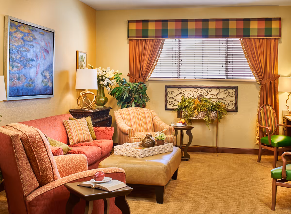A cozy living room with a red patterned armchair and sofa, a striped armchair, and a beige ottoman in the center. The room features a side table with a lamp and flowers, a window with orange curtains and a colorful valance, and decorative wall art. There are also green plants and a small table with a cup and fruit.