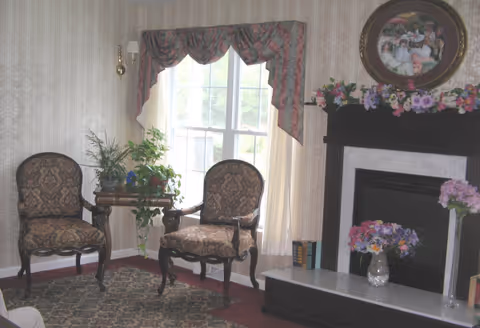 Two upholstered armchairs flank a small plant-covered table in front of a window beside a mantel decorated with flowers and a framed picture.